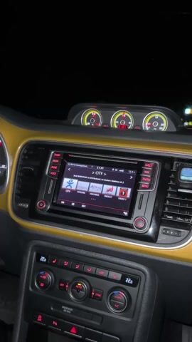 The video shows the interior of a car, likely a Volkswagen Beetle, driving at night. The dashboard, finished in a distinct gold or yellow tone, is the focal point. A built-in infotainment system is displaying a radio station interface with the station "CITY" selected. Above the central console, three gauges are visible, adding to the car's retro aesthetic. The road ahead is visible through the windshield, illuminated by streetlights and the headlights of other cars, indicating the car is traveling on a highway or similar roadway. The interior lighting is minimal, creating a dark ambiance which serves to highlight the dashboard and road ahead. The car seems to be traveling at a moderate speed.