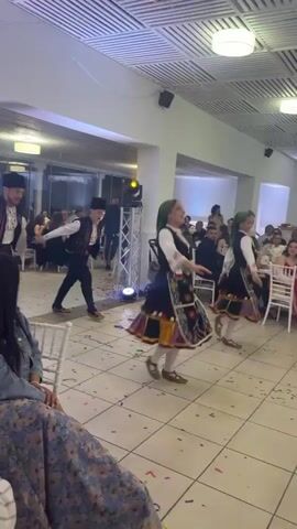 Inside a brightly lit hall, a group of four people—two men and two women—are performing a traditional folk dance. They are dressed in what appears to be traditional attire. Confetti litters the tile floor around them, suggesting a celebratory event. Around them, people are seated at tables, observing the dance. A baby stroller is also visible. The hall has a grid-patterned ceiling and large windows. A woman in a floral dress is seated in the foreground, partially obscuring the view of the dancers.