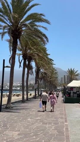 This media file captures a lively scene along a sun-drenched promenade lined with tall palm trees. The pathway, paved with irregular stones, leads towards a distant town nestled against a backdrop of hazy mountains under a clear blue sky. The time of day appears to be midday, indicated by the bright sunlight casting distinct shadows on the ground.

In the foreground, a couple walks hand-in-hand, the woman in a pink dress and the man in a light-colored shirt and shorts, each carrying a white shopping bag. They are walking away from the camera, towards the other pedestrians on the promenade. Behind them, to the left, is a beach with lounge chairs and umbrellas, suggesting a resort or tourist area. To the right of the promenade, a white tent or awning is visible, possibly part of a market or food stall, with people milling around it.

Further down the promenade, a steady stream of people are seen walking in both directions, indicating a popular spot. The overall atmosphere is one of leisure and enjoyment, with the bright sunshine and the presence of beachgoers and shoppers contributing to a summery, vacation-like ambiance. The location is identified as Arona, Spain, which aligns with the visual cues of palm trees, a sunny coastal setting, and mountainous terrain. No visible text is present in the media.