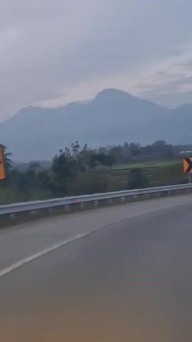 The video captures a view from a vehicle traveling on a road, likely a highway. In the foreground, the asphalt road curves to the right. A silver guardrail runs alongside the road, with red reflectors. Beyond the guardrail, a green field is visible, suggesting a rural or agricultural area. In the mid-ground, a yellow diamond-shaped road sign with a black chevron indicating a curve to the right is prominently displayed. In the background, several mountain ranges are visible, shrouded in a hazy, pale blue atmosphere, with a cloudy sky overhead. The overall lighting suggests it is either dawn or dusk. There are no people or animals visible in the frame.