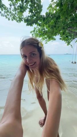 A young woman, seen from a low angle, smiles into the camera at the start of a short video. She has long blonde hair and wears a white bikini and a white flower over her ear. She stands on a white sand beach in front of clear, turquoise water. In the distance, a boat is visible on the horizon. The camera is held at arm’s length, obscuring part of the view. The woman then stands upright, and walks into the water. She turns and raises her hands overhead. A tree with green leaves provides shade, and a swing hangs from one of its branches.