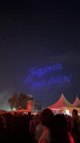 The video shows a large crowd of people at an outdoor event, likely a concert or festival, under a dark sky at night. In the beginning, there are drones in the sky forming words, possibly in Spanish. Then, the scene shifts to a stage area with lights and smoke, suggesting a performance. People are standing and watching the show, with some holding up their phones to record. The environment appears to be an open space with some structures nearby.