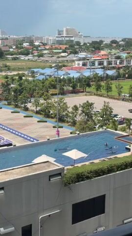 The video shows an aerial view of an outdoor swimming pool on a rooftop. Several people are visible in the pool, some of whom appear to be swimming or floating. Two white umbrellas are partially submerged in the water. The pool is surrounded by greenery and overlooks a city with buildings, trees, and a parking lot. The sky is overcast with a hint of rain in the distance. The camera is positioned slightly above the pool, looking downwards.