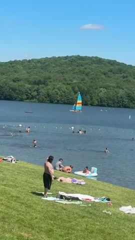 The video captures a sunny day at a lake with numerous people enjoying recreational activities. In the foreground, a vibrant green grassy bank is dotted with individuals sitting, lying on towels, standing, and changing clothes. Several people are seen in the water, swimming, splashing, and floating on inflatable rafts. A colorful sailboat, with yellow, red, and blue sails, is visible in the middle distance of the lake during the initial frames. A line of buoys marks a designated swimming area in the water. The background features lush green hills densely covered with trees, all under a clear blue sky with sparse white clouds.