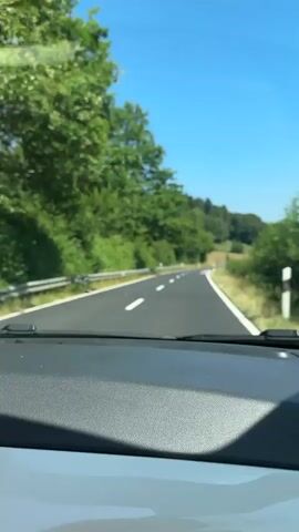 The video shows a view from inside a vehicle driving on a paved road through a wooded area. Trees and green foliage line both sides of the road. The sky is clear and blue. A side mirror reflects the road behind the car. The final shot shows a blonde woman with sunglasses in the passenger seat, her hair blowing in the wind, smiling.