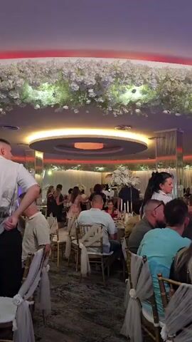 The video shows an indoor event, possibly a wedding reception or a formal dinner. The room is decorated with an abundance of white floral arrangements hanging from the ceiling and along a central circular light fixture that emits a reddish glow. Tables are set with chairs draped in white fabric, and guests are seated or standing around them. In the foreground, a person in a white shirt with a graphic print walks away from the camera. To their right, a woman wearing a black apron over a white shirt and white gloves walks towards the camera, holding a bottle and a stopper. Several other guests are visible in the background, some dancing or conversing. The lighting is warm and ambient, contributing to a celebratory atmosphere.