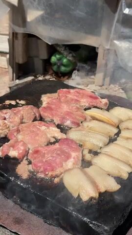 The video shows thinly sliced raw meat and pieces of fatty pork belly being cooked on a hot flat stone grill. Flames are visible to the left of the grill, indicating a heat source. Smoke rises from the food as it cooks. In the background, obscured by a plastic sheeting and wooden structure, is a green object, possibly a bell pepper, placed near the heat source. The overall setting appears to be outdoors, with visible dirt and loose debris on the ground. The footage is filmed from a slightly elevated, close-up perspective.