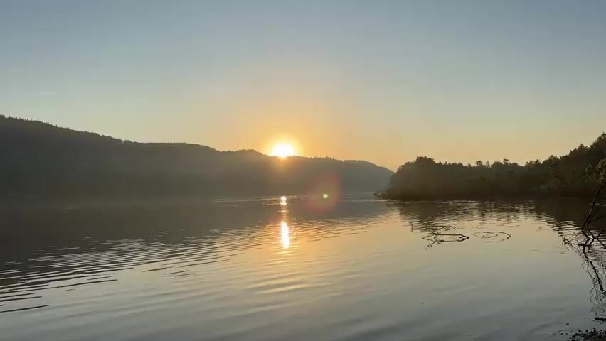 The video shows a serene sunrise over a body of water. The sun is just cresting the hills in the background, casting a warm glow on the water and the surrounding landscape. Gentle ripples spread across the surface of the water, reflecting the sunlight. The hills on either side of the water are covered in trees, and a faint mist hangs in the air. The sky is a clear blue, with a few wispy clouds. The overall mood of the video is peaceful and tranquil.