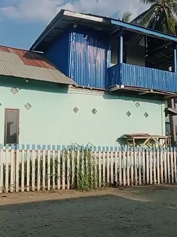 The video shows a light blue two-story house with a blue corrugated metal upper story and balcony. A white picket fence with blue trim runs in front of the house, separating it from a dirt road. Tall green grass grows behind the fence. Palm trees are visible in the background. The sky is blue with some clouds.
