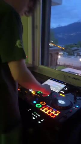 A DJ plays music on equipment in front of a window. The view outside the window shows a landscaped backyard with a pool, surrounded by grass and decorative lighting. Beyond the backyard, there are houses and trees, with a backdrop of snow-capped mountains under a blue, cloudy sky. The DJ is partially visible, focusing on the mixing equipment with illuminated buttons and a screen. The video appears to be taken during dusk or early evening, evidenced by the lighting and the color of the sky.