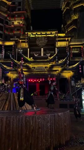 Three performers dance on a wooden stage, holding props that appear to be chains or ropes. The stage is situated in front of an ornately lit building with traditional East Asian architectural features. Other performers are also visible in the background. The area surrounding the stage is paved, and the lighting creates a vibrant, colorful atmosphere. The scene appears to be part of a cultural performance or theatrical production, set against the backdrop of traditional architecture and bright lights.