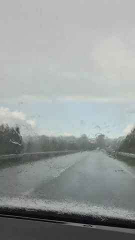 The video shows a view from inside a vehicle driving on a wet road. Rain is visible on the windshield and the windshield wipers are clearing the view periodically. The road appears to be a two-lane highway with trees and vegetation on either side. The sky is overcast and the lighting suggests it is daytime. The overall scene depicts driving in rainy conditions.