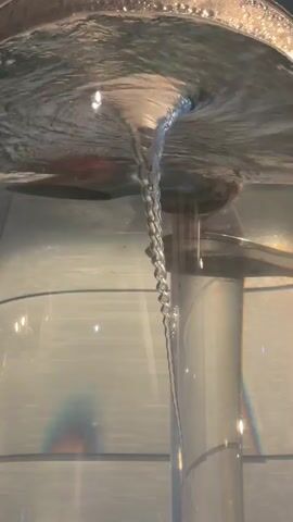 A close-up shot shows a vortex of water in a clear container. The vortex is a narrow column of water that appears to be moving downwards. The water is clear, and the vortex is visible against a background of blurry lights and shapes. The lighting in the scene is bright, creating reflections on the surface of the water. The shot appears to be taken from slightly below the surface of the water.