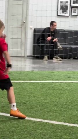 A young boy in a red soccer jersey and black shorts is playing soccer on an indoor turf field. He is wearing orange soccer cleats and white socks. He appears to be looking to his right. In the background, there are spectators sitting on couches and chairs behind a net. The environment is an indoor sports facility.