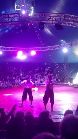 Two performers in circus attire are on a brightly lit pink stage under a tent. The performer on the left is wearing a white shirt and dark pants and is holding a white bucket. The performer on the right is wearing a white shirt and suspenders with dark pants and is holding a long stick. There is a large audience seated in the background. The stage is illuminated with purple and pink lights.