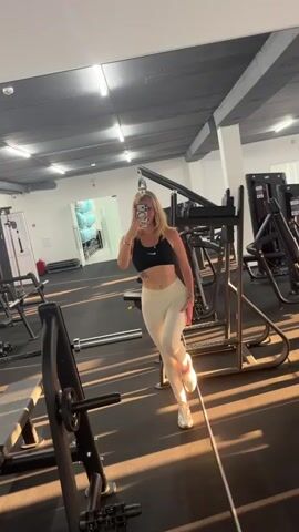 The video shows a young blonde woman in athletic wear performing a series of leg stretches in a gym. She is standing in front of a mirror and the camera is positioned in front of her, capturing her reflection and the gym environment. The gym appears to be equipped with various weight machines and other fitness equipment. The lighting in the gym is artificial and there is sunlight casting long shadows across the floor. The woman's movements are fluid and controlled as she demonstrates different stretching poses. She is holding her phone to take a selfie or record herself. Her attire consists of a black sports bra and light-colored leggings. The background shows gym equipment, including weight racks and benches. The overall impression is of a workout session focused on flexibility.