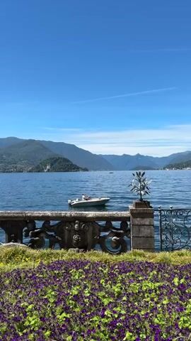 A picturesque landscape scene unfolds, showcasing a vast, calm body of water reflecting a clear, bright blue sky. Rolling green hills and mountains form a scenic backdrop across the water. In the foreground, a decorative stone balustrade with ornate carvings and finials separates a lush, vibrant garden from the water. The garden is filled with a profusion of purple and yellow flowers, interspersed with green foliage. Beyond the balustrade, a small white boat with two occupants is visible on the water, gliding gently. To the left of the frame, a large palm tree stands prominently, adding a tropical element to the setting. Two distinctive, twisted stone columns with square tops are positioned near the balustrade, adding architectural interest to the scene. The overall impression is one of serene beauty and idyllic tranquility.