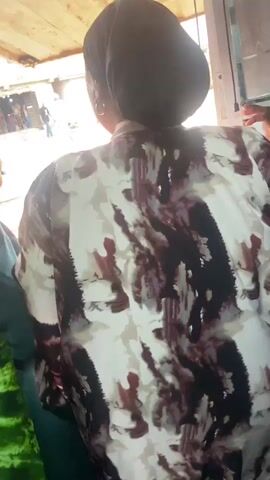 The video shows three people standing near a window overlooking a street. One person wears a green tie-dye patterned garment, another wears a white and brown patterned shirt, and the third wears a dark colored top.  The people are looking out the window at the street. The street is visible in the background with people walking.