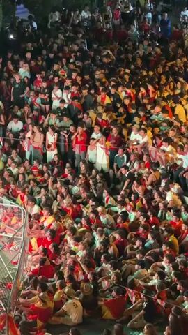 A dense crowd of people, mostly young adults, are gathered outdoors at night. They are wearing red and yellow clothing, suggesting they are supporting a team or participating in an event. Some individuals are holding flags and banners. The crowd appears to be seated and looking in one direction, likely at a performance or event. The lighting is dim, with some artificial lights illuminating the scene.