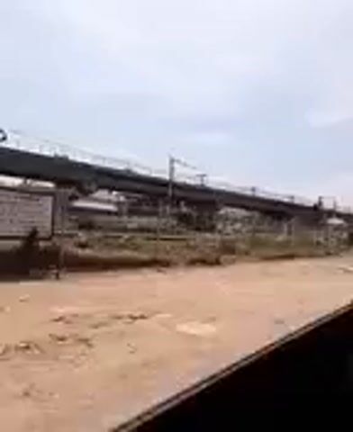 The view from inside a vehicle, possibly a car, shows a partially constructed elevated structure with a railroad crossing and traffic lights visible outside. The ground appears to be dirt. The sky is overcast. The video seems to be taken from the passenger seat of a moving vehicle. A person’s hand is briefly visible inside the car.