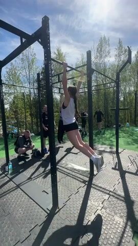 Here is a description of the video:

The video shows a group of people exercising outdoors on a sunny day. The central focus is a woman doing pull-ups on a black metal rig. She is wearing a white tank top and black shorts. Several other people are nearby, some watching and some participating in similar exercises. The ground is covered with gray interlocking mats, and there is a patch of green artificial turf visible in the background. Trees surround the area, and the sky is blue with some clouds.