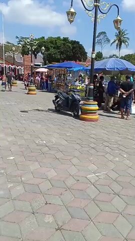 A woman wearing a pink shirt and a black hijab walks with a young girl in a red outfit. They are on a paved walkway in an outdoor market. The walkway is made of grey and red patterned tiles. There are stalls set up with various items for sale. One stall has a menu with pictures of food. Several people are browsing at the stalls. A motorcycle is parked near one of the stalls. The sky is blue and there are trees in the background.