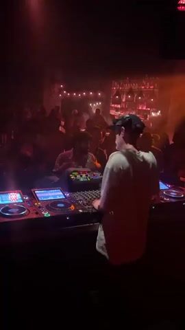 A DJ stands behind a console in a dimly lit club, wearing a white t-shirt and a baseball cap. The console has multiple screens and buttons, with two turntables on either side. The DJ appears to be operating the equipment. In the background, a crowd of people is visible, some standing and some sitting. The environment is a nightclub with red and orange lighting, and decorative lights hanging from the ceiling. The overall atmosphere suggests a music performance or party.