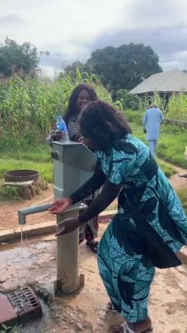 Two women are by a water pump in a rural setting. One woman in a patterned dress is pumping water with her hands. Another woman in a similar patterned dress is holding a water bottle. A man in a blue outfit walks away from the scene in the background, where there are corn stalks and a building. The ground around the pump is wet and dirty, with a metal grate to the left. The sky is cloudy.