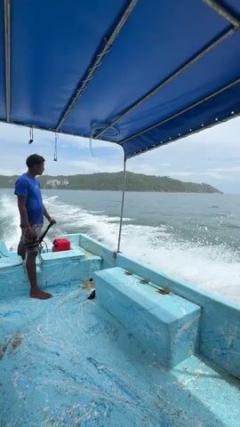 A medium shot from inside a boat captures a person standing on the left, holding the boat's tiller, and looking toward the camera. The person, a light-skinned male with dark hair, wears a blue short-sleeved shirt and floral-patterned shorts. He is barefoot. The boat's floor is blue and white with paint splatters. The boat has blue wooden benches, and a small red fuel tank is visible on the floor behind the person. A blue canopy is overhead. The boat is in motion, creating a white wake behind it. The water is a dark blue, and in the distance, a tree-covered landmass is visible under a light blue sky with some clouds.