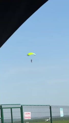 A person is parachuting down from the sky. They are attached to a yellow and blue parachute. They are descending towards a grassy field enclosed by a green fence. A small airplane is visible in the distance behind the fence. A sign in Russian that says "Smoking is Prohibited" is attached to the fence. The sky is clear and blue. The video is shot from a vehicle, with the frame of the car window visible at the top.