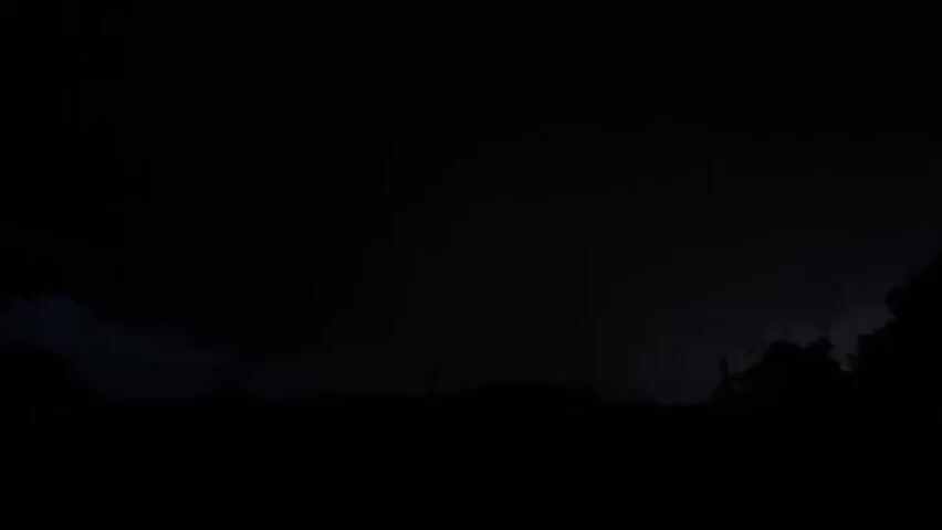 The video shows a dark, silhouetted landscape at night, likely a rural or suburban area with trees on the right and some structures in the middle ground. The sky is filled with ominous, dark clouds. Lightning flashes intermittently across the sky, illuminating the clouds and the landscape in brief, bright bursts. The lightning appears to be originating from within the cloud formations and striking both within the clouds and possibly towards the ground. The overall mood is dramatic and turbulent due to the storm.