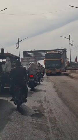 A vertical, medium-shot, eye-level perspective video captures a traffic jam on a bridge under a cloudy sky. In the foreground, a motorcycle with a rider is stopped on the right side of the road. To the left, the back of a truck with people in the cargo bed is visible. Further ahead, a line of vehicles including motorcycles, a truck, and a yellow double-decker bus are stopped. The bridge has streetlights and wires overhead, and a red flag is visible on the right side of the road in the background. The road surface is asphalt and appears wet in patches.