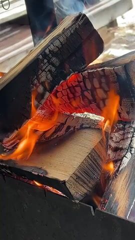 A close-up shows a fire burning inside a metal container, with flames engulfing several pieces of wood. The wood shows charring and patterns. Some smoke is visible. The background includes a metal grill rack and glimpses of a person's legs and dark clothing. The focus is primarily on the burning wood and the intense orange flames.