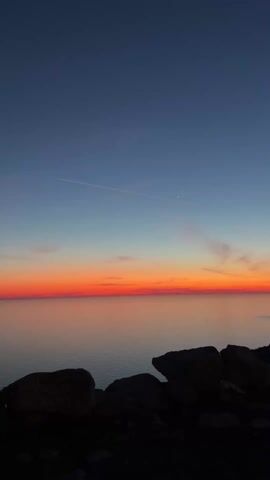 The video shows a sunset over the ocean. The sky is dark blue at the top, fading to orange and red near the horizon. There are a few small clouds in the sky, and a contrail from an airplane. The ocean is calm and reflects the colors of the sky. In the foreground are dark rocks that line the coast. A person's voice can be heard in the background.