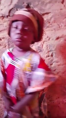 A young child wearing a colorful, patterned robe and a brown hat stands in front of a rough, beige wall. The child looks towards the camera and appears to be speaking or singing, though the audio is unclear. The child makes small hand movements and shifts their weight slightly. The lighting is somewhat dim and has a slightly reddish tint. The background wall has a textured appearance, resembling cracked or weathered stone.