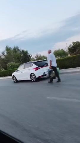 A group of people are gathered around several parked cars. The video appears to be filmed from a moving vehicle, as the perspective shifts and blurs. The environment is outdoors, with trees and a cloudy sky visible. Some of the cars appear to be modified or show signs of wear. The people are casually dressed and some are interacting with each other or the cars. The overall atmosphere seems relaxed, possibly a car meet or gathering.