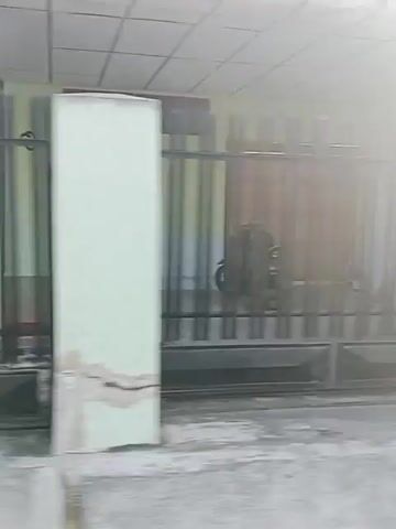 A handheld, panning video shows an outdoor scene. The environment appears to be a somewhat cluttered marketplace or street with a few small buildings or stalls. In the foreground, there's a large dark pot. The camera pans to the right, showing a simple wooden structure, possibly a stall, with some red elements on top. Further to the right, a wall with graffiti is visible. The camera continues to pan, revealing a motorcycle parked on the left side. The scene then shifts to a building with red double doors. The lighting suggests it is daytime, possibly late afternoon, with bright sunlight causing some overexposure. There are no people visible in the video.