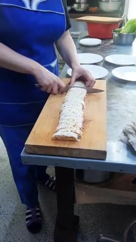A person wearing a blue apron is slicing a long, rolled food item on a wooden cutting board. The food item appears to be a wrap or shawarma, cut into several thin, circular slices. The cutting board is placed on a metal table. Several white plates are visible in the background, along with kitchen appliances and a bowl of lettuce. The person is using a knife to make the slices. The lighting suggests an indoor kitchen environment.