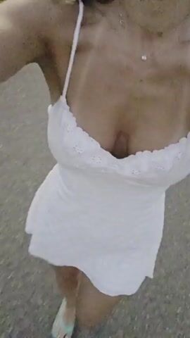 A first-person perspective video shows a person walking on a paved surface. They are wearing a white sundress with thin straps and a ruffled neckline, and flip-flops. The person is moving forward, with their legs and feet visible. The background is a blurred outdoor setting, suggesting they are walking on a sidewalk or path. The lighting indicates it is daytime, possibly late afternoon or early evening, with warm tones. The video appears to be a casual, personal recording, capturing a simple moment of movement. No text is visible.