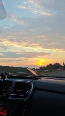 The video shows the view from inside a car dashboard. Outside the car, a sunset is visible in the sky with clouds. There are trees and a building visible on the side of the road. The car appears to be moving on a road.