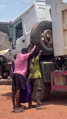 Three men are struggling to lift a large tire onto the side of a white dump truck. The men are dressed casually in t-shirts, shorts, pants, and sandals. The truck is parked on a dirt or gravel surface, and other similar trucks are visible in the background. The sky is blue with some clouds. It appears to be a daytime scene.