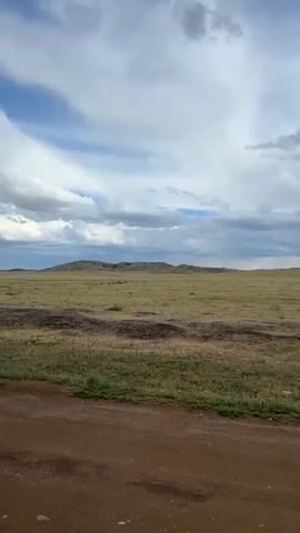 The video captures a vast, open grassland under a dynamic sky. The foreground is dominated by a dirt road, its reddish-brown surface textured with tire tracks, suggesting recent vehicle passage. To the right of the road, a strip of green grass and some dry vegetation lines the edge.

Beyond the road, the landscape unfolds into rolling hills covered in dry, golden grass, interspersed with patches of greener vegetation. The hills are gently sloped and stretch towards a distant horizon. In the far distance, a faint outline of more substantial hills or a mesa can be seen.

The sky is a dramatic display of contrasting clouds. Bright, white cumulus clouds are scattered across a bright blue sky, while darker, heavier storm clouds gather in the distance, hinting at potential rain or a coming storm. Shafts of sunlight pierce through the clouds in places, illuminating sections of the grassland.

The time of day appears to be late afternoon or early evening, given the quality of the light and the cloud formations. The overall mood is one of natural beauty and vastness, perhaps with a touch of dramatic anticipation due to the weather. There are no people or distinct objects visible in the scene, emphasizing the natural, undeveloped setting. The location is identified as Bakty, Kazakhstan, which aligns with the image of a vast, arid grassland. There is no visible text in the video.