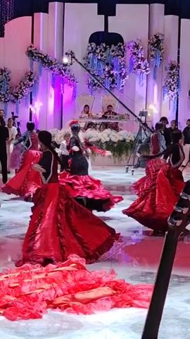 A group of female dancers in vibrant red flamenco dresses perform on a stage illuminated by purple and white lights. The dancers are positioned throughout the frame, with some closer to the camera and others further back. In the background, a couple is seated at a table, presumably honored guests or newlyweds, behind a floral arrangement. Several individuals are observing the performance, and film equipment, including camera booms, is visible, suggesting the event is being professionally recorded. The stage floor is reflective, creating a mirror-like surface that reflects the lights and movement. The overall atmosphere appears festive and celebratory, likely at a wedding or similar formal event.