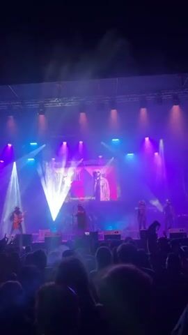 The video shows a band called Nine Mile performing on a stage in front of a crowd. There are five visible band members, including a singer, guitarist, and drummer.  The stage is brightly lit with purple and blue lights and smoke. Behind the band is a large screen with the band's name and an image of what appears to be Bob Marley. The crowd is densely packed and appears to be enjoying the show.