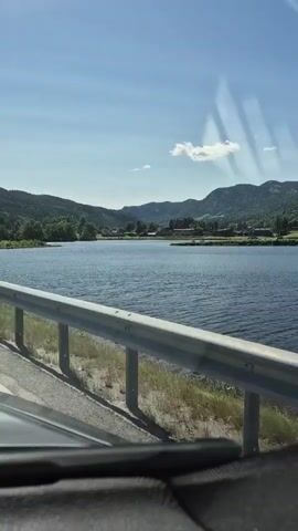 The video shows a view from a moving vehicle looking out over a body of water. In the foreground, a metal guardrail runs along the side of a road, with the edge of the vehicle visible at the bottom. A road sign is positioned on a pole to the left of the frame. Beyond the guardrail, a large expanse of blue water stretches out, reflecting the clear sky above. In the background, rolling hills covered in green trees and some buildings are visible. The sky is a bright blue with a few scattered white clouds. The sun appears to be shining, casting shadows and creating reflections on the water. The overall impression is one of a scenic drive through a natural landscape.