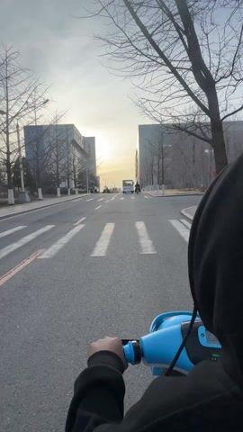 The video shows a first-person perspective of someone riding a blue scooter on a paved road. The road has a crosswalk painted on it, and there are modern buildings on either side. The buildings have a glass and metal exterior and are connected by a sky bridge. Bare trees line the road, and the sky is a mix of grey and yellow, indicating either sunrise or sunset. Other vehicles, including a white van and a scooter, are visible in the distance. The person riding the scooter is wearing a black hooded jacket and holding the scooter's handlebars.