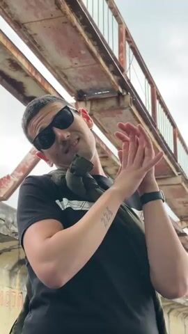 A man with dark hair and sunglasses smiles and makes hand gestures toward the camera, under an iron bridge. He wears a black t-shirt with a white stripe across the chest and a green bag slung over his shoulder. He has a tattoo on his left forearm. The bridge overhead is made of metal beams and is rusty in appearance. The sky is overcast.