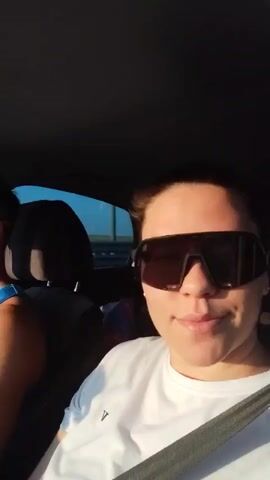 The video shows two people in the front seats of a car, driving on a highway. The woman is in the passenger seat and is looking directly at the camera. She is wearing a white t-shirt and large sunglasses. The man is driving. He is wearing a blue tank top and sunglasses. He is looking at the road.  Sunlight is streaming into the car from the side. A highway guardrail is visible out the window.  Music is playing in the background.