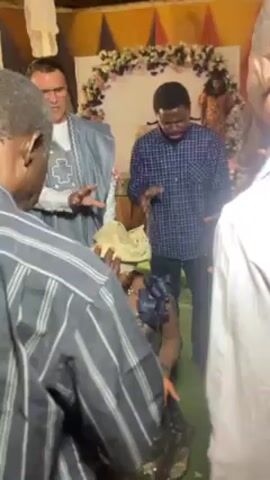 The video shows a group of people gathered indoors, appearing to be praying or performing a blessing. A woman is kneeling, and several people stand around her with their hands extended towards her. One man, wearing a light blue robe, is making gestures with his hands while speaking. Another man stands next to him in a blue and white checkered shirt. The background includes a floral archway and draped white fabric. The room seems to be decorated for an event.