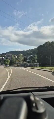 The video shows a view from inside a vehicle, looking forward. The vehicle is on a paved road with white lane markings. There are traffic signals and street signs visible. To the left, there is a blue car turning. In the background, there are houses built on a hillside, surrounded by trees. The sky is blue with white clouds. The footage is shaky, indicating movement of the vehicle.